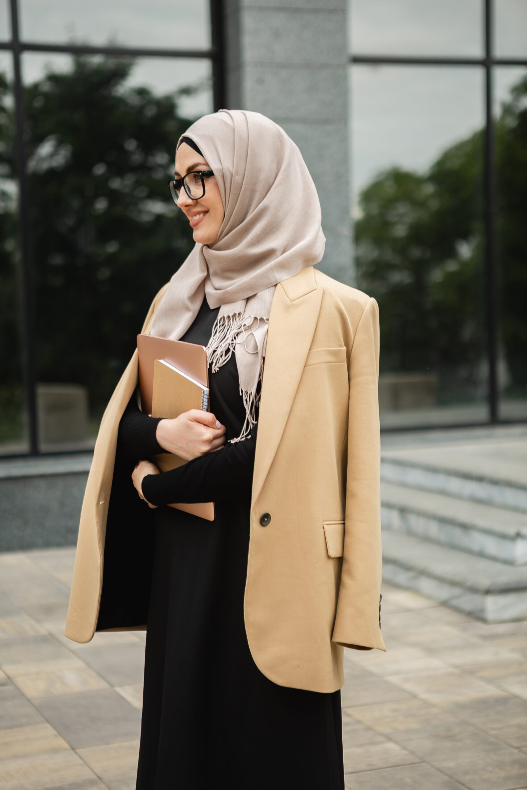 modern stylish muslim woman in hijab, business style jacket and black abaya walking in city street with laptop, freelancer online remote work, business woman in glasses